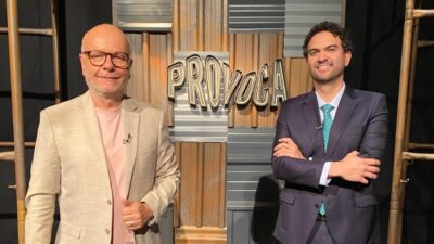 Marcelo Tas de blazer claro e Felipe Nunes de terno escuro posam sorrindo diante do letreiro Provoca
