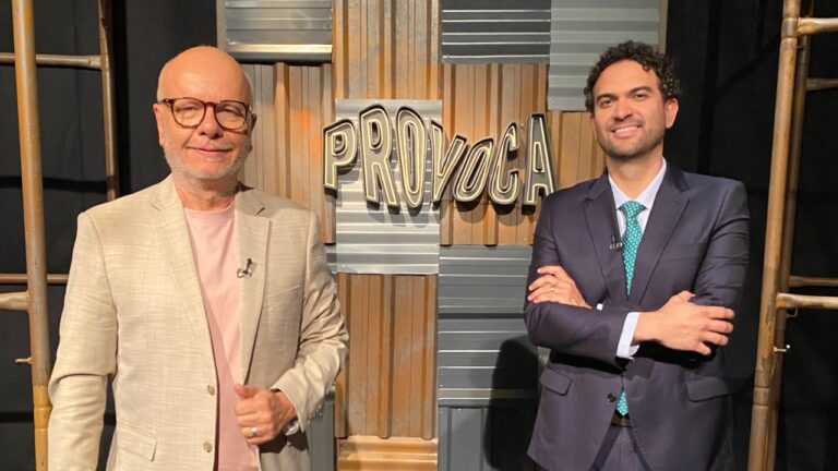 Marcelo Tas de blazer claro e Felipe Nunes de terno escuro posam sorrindo diante do letreiro Provoca