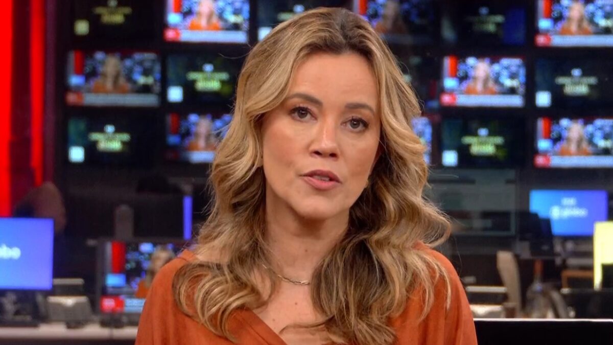 Natuza Nery de blusa laranja fala diante das câmeras em redação da GloboNews com diversos monitores ao fundo