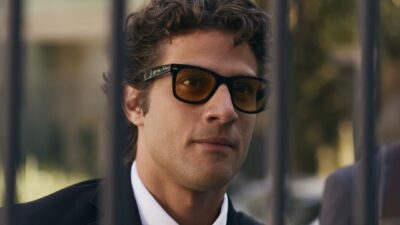 Pedro (Chay Suede) de terno e óculos escuros, com expressão séria, diante de grades em ambiente externo