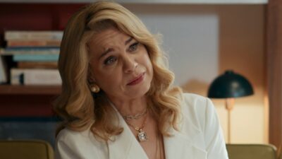 Pilar (Isabel Teixeira) de blazer branco inclina a cabeça enquanto conversa em escritório na novela Quem Ama Cuida