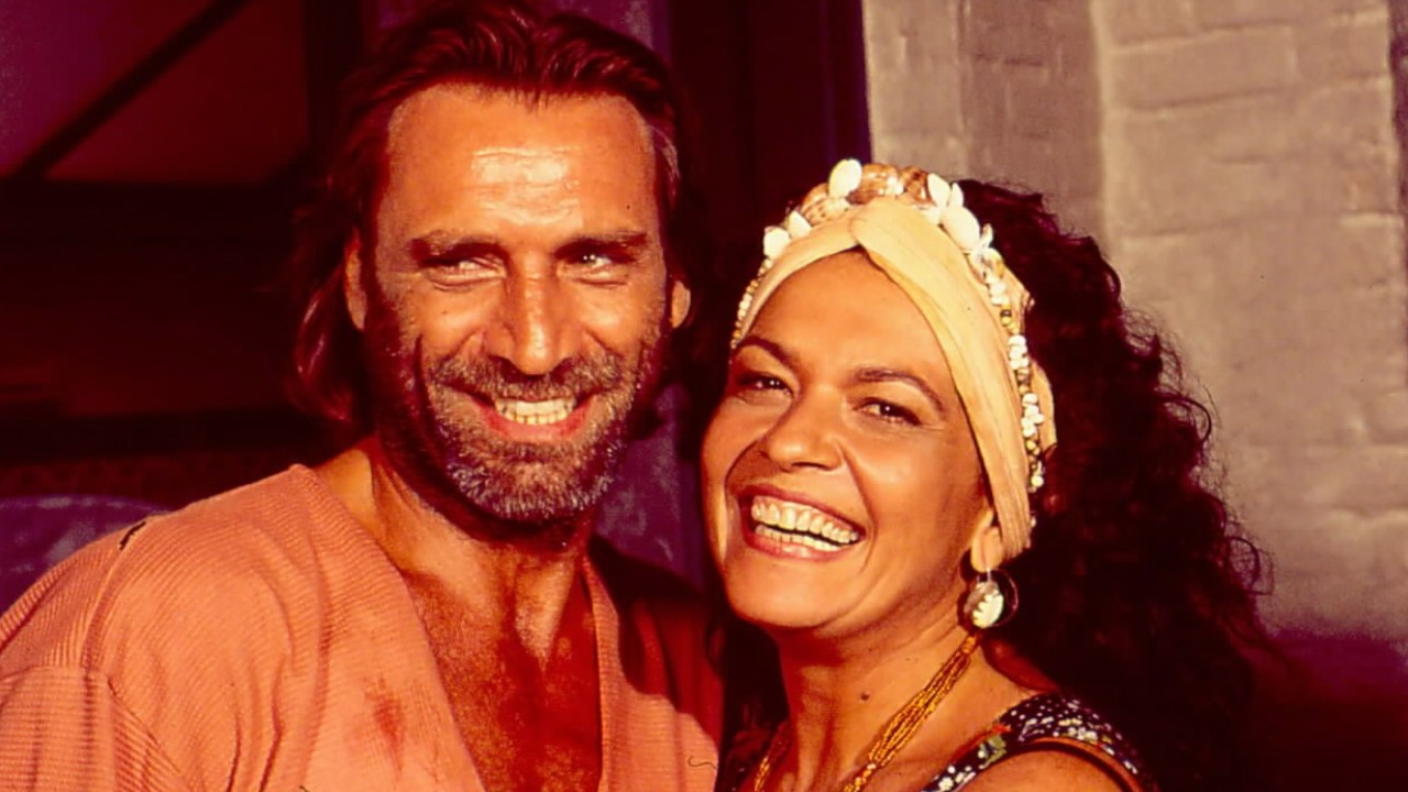 Ramiro (Herson Capri) e Serena (Regina Dourado) sorriem juntos; ela usa turbante com búzios e brincos grandes