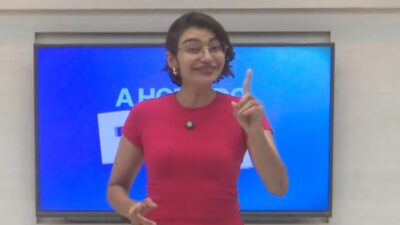 Soraya Almeida de blusa vermelha sorri e ergue o dedo indicador diante de uma tela azul em estúdio