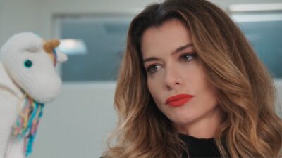 Vanessa (Alinne Moraes) de cabelo ondulado e batom vermelho olha para o lado ao lado de um unicórnio de pelúcia