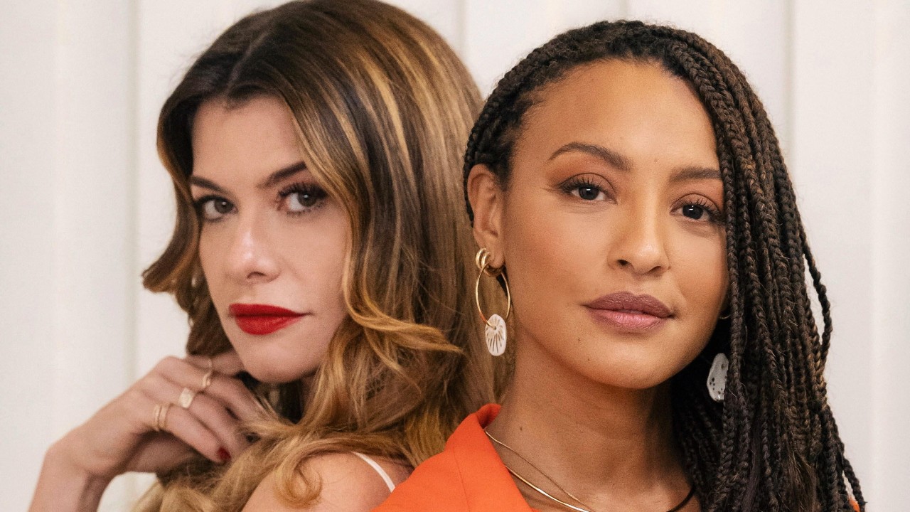 Vanessa (Alinne Moraes) com cabelo ondulado posa junto a Bela (Sheron Menezzes), de tranças e blazer laranja