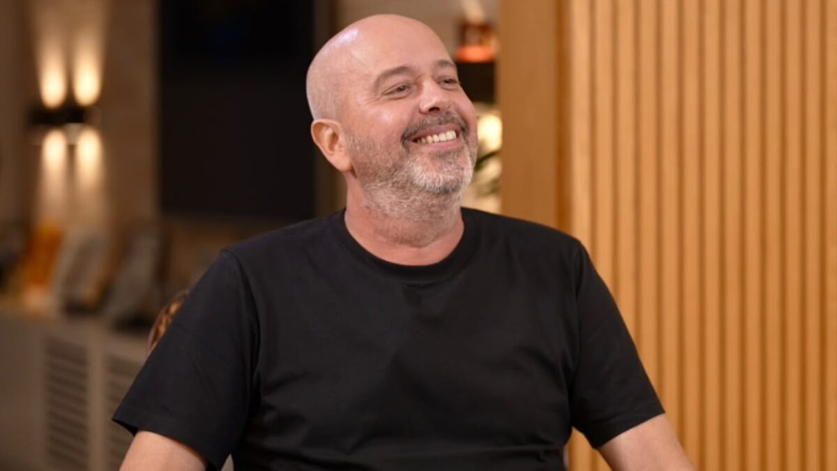 Alex Escobar de camiseta preta sorri sentado em ambiente interno com painel de madeira ao fundo