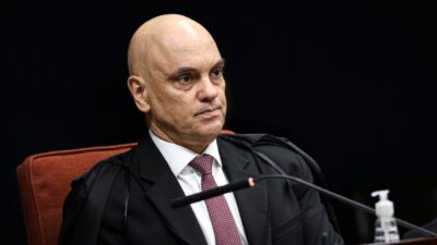 Alexandre de Moraes de toga e gravata vermelha está sentado diante de microfone, com expressão séria e fundo escuro