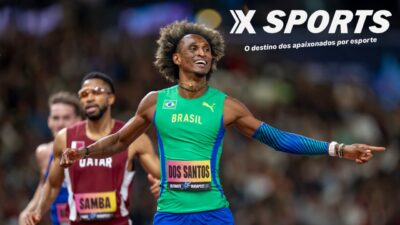 Alison dos Santos sorri com uniforme verde do Brasil e braços abertos após prova de atletismo, com adversários ao fundo
