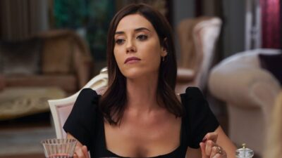 Asya (Cansu Dere) de vestido preto sentada à mesa com expressão séria em cena da novela turca Iludida