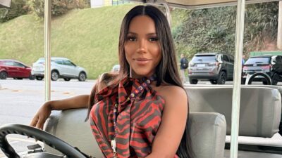 Carol Lekker sorri sentada ao volante de um carrinho, com blusa estampada vermelha e preta e carros ao fundo