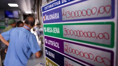 Painel em casa lotérica exibe prêmios da +Milionária, Mega-Sena e Lotofácil, com clientes ao fundo