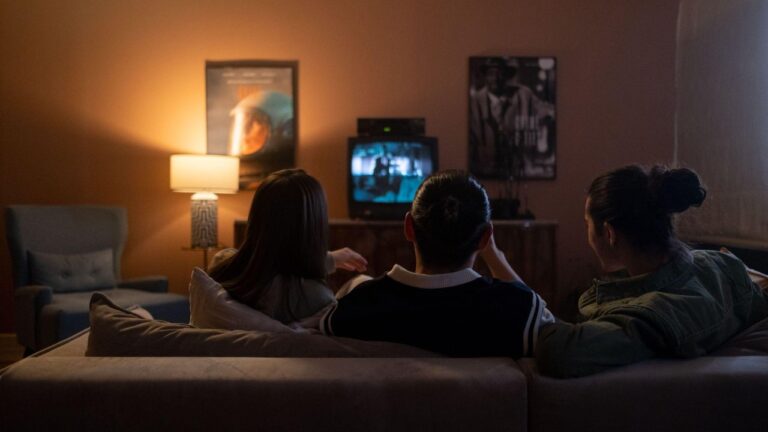 Três pessoas sentadas em um sofá assistem à televisão em uma sala com iluminação baixa