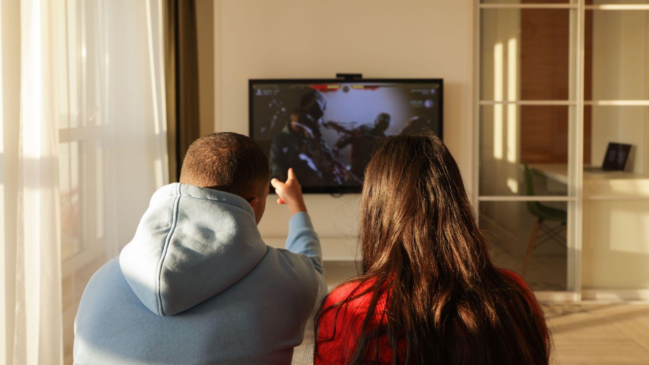 Duas pessoas sentadas de costas assistem a um programa exibido em uma televisão na sala