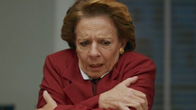 Diná (Rosi Campos) de casaco vermelho cruza os braços e olha para baixo com expressão tensa em Quem Ama Cuida