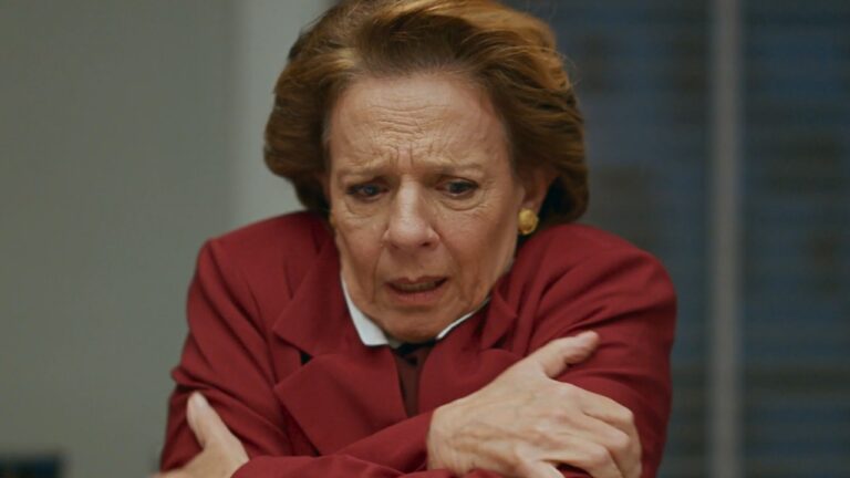 Diná (Rosi Campos) de casaco vermelho cruza os braços e olha para baixo com expressão tensa em Quem Ama Cuida