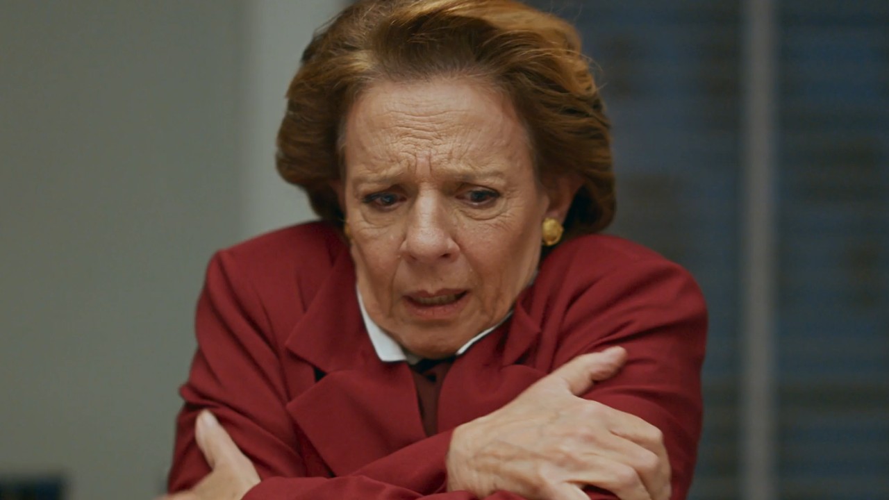 Diná (Rosi Campos) de casaco vermelho cruza os braços e olha para baixo com expressão tensa em Quem Ama Cuida