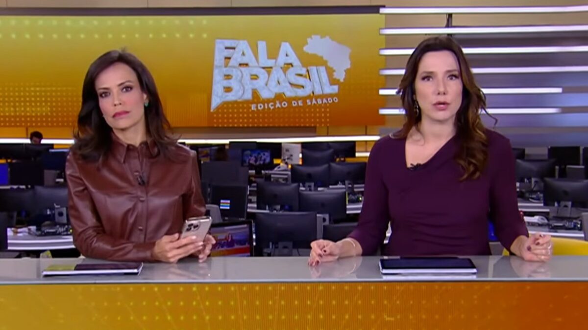 Fabiana Oliveira segura celular ao lado de Adriana Perroni na bancada do Fala Brasil, com redação ao fundo