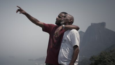 Dois homens abraçados observam a paisagem enquanto um deles aponta para o horizonte com montanhas ao fundo