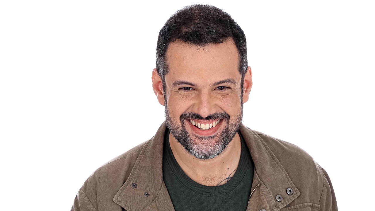 Gui Alves sorri de jaqueta marrom e camiseta verde, com barba curta, diante de fundo branco em Herança em Jogo