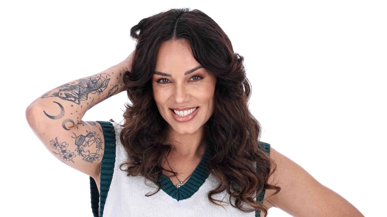 Jamile Salomão sorri com braço tatuado atrás da cabeça, cabelo longo e blusa branca e verde diante de fundo branco