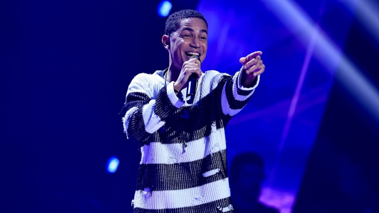Junior Azevedo canta ao microfone e aponta para a plateia durante as Audições às Cegas do The Voice Brasil