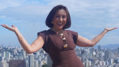 Katiuzia Rios sorri com os braços abertos, usa vestido marrom e colar dourado, com prédios da cidade ao fundo