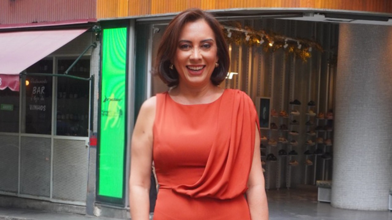 Katiuzia Rios sorri com vestido laranja sem mangas diante de lojas em uma rua comercial
