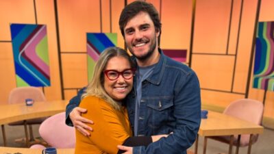 Katy Navarro de óculos vermelhos abraça Hugo Bonemer, de jaqueta jeans, no estúdio do Sem Censura