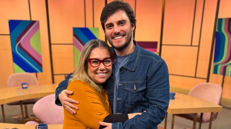 Katy Navarro de óculos vermelhos abraça Hugo Bonemer, de jaqueta jeans, no estúdio do Sem Censura