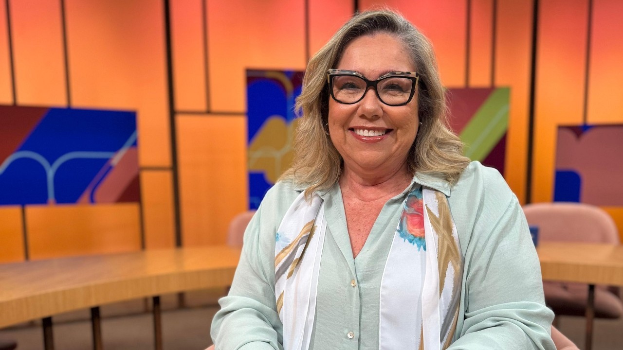 Katy Navarro sorri de óculos, camisa verde-clara e lenço estampado em estúdio com painéis coloridos e mesas ao fundo