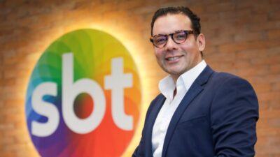 Klayton Cavalcante de terno azul e óculos sorri diante do logotipo do SBT