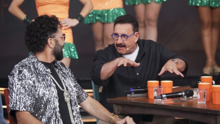 Latino de camisa prateada ouve Ratinho, de óculos azuis, gesticular diante de mesa com microfones e canecas