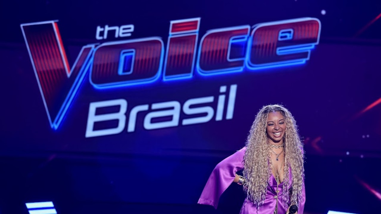 Lia Lira sorri no palco diante do logotipo do The Voice Brasil após apresentação nas Audições às Cegas