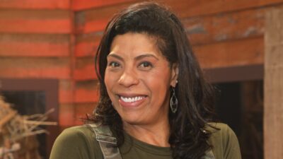 Márcia Fu sorri de blusa verde e brincos longos, com cabelo preto solto e cenário em tons de madeira ao fundo