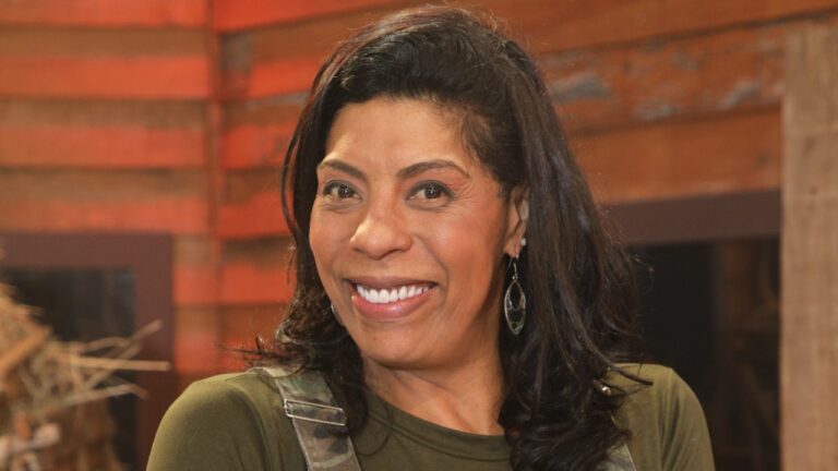 Márcia Fu sorri de blusa verde e brincos longos, com cabelo preto solto e cenário em tons de madeira ao fundo