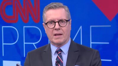 Márcio Gomes de terno e gravata fala diante de painel azul com logotipo da CNN e identificação do CNN Prime Time
