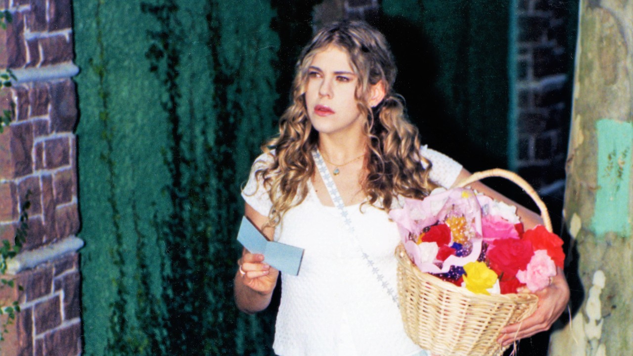 Marisol (Bárbara Paz) segura cesta de flores e um cartão diante de uma casa