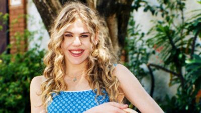 Marisol (Bárbara Paz) sorri com vestido azul estampado em área externa com árvores ao fundo