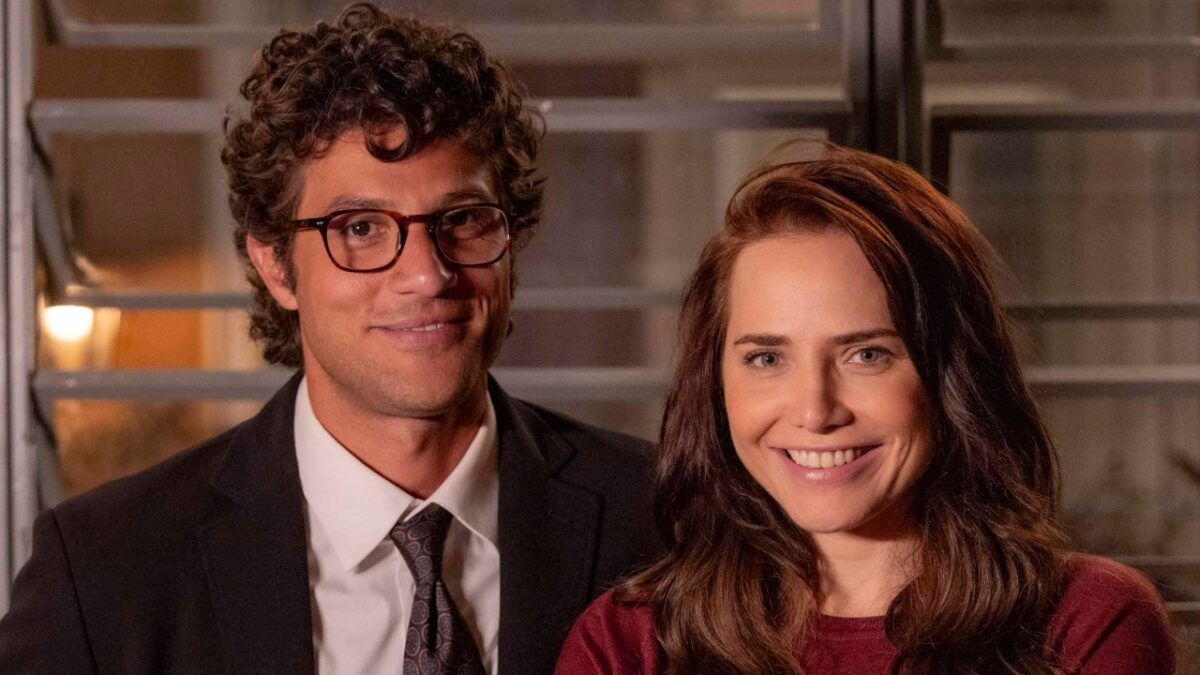 Pedro (Chay Suede) de terno e Adriana (Leticia Colin) de blusa vinho sorriem lado a lado em Quem Ama Cuida