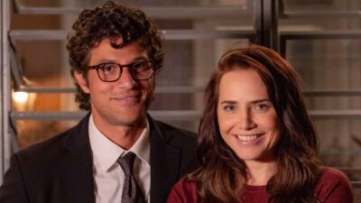 Pedro (Chay Suede) de terno e Adriana (Leticia Colin) de blusa vinho sorriem lado a lado em Quem Ama Cuida