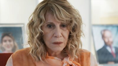 Pérola (Renata Sorrah) de blusa laranja olha para baixo, com retratos desfocados ao fundo em Por Você