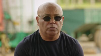 Rick Sollo careca, de óculos escuros e camiseta preta, sentado diante de ambiente externo desfocado