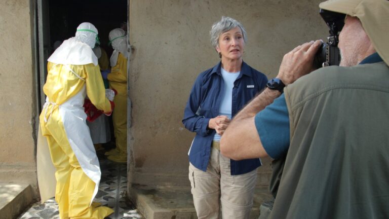 Sônia Bridi fala diante da câmera enquanto profissionais com trajes de proteção trabalham ao fundo no Congo