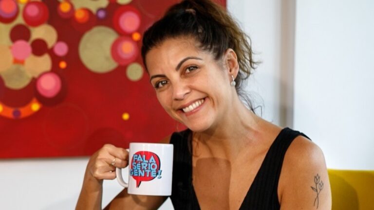Thalita Rebouças sorri segurando caneca do Fala Sério, Gente!, de blusa preta e cabelo preso, com quadro vermelho ao fundo
