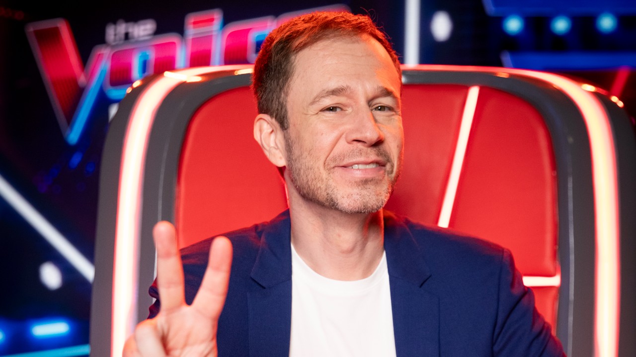 Tiago Leifert sorri e faz sinal de V diante da cadeira vermelha do The Voice Brasil