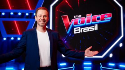 Tiago Leifert de blazer azul e camiseta branca sorri de braços abertos diante do logo do The Voice Brasil