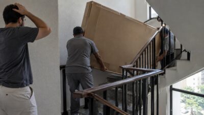 Dois entregadores carregam uma grande caixa de TV pela escada de um prédio enquanto um homem observa a dificuldade na passagem
