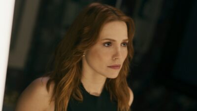 Adriana (Leticia Colin) aparece com cabelos ruivos soltos e blusa preta sem mangas, olhando atentamente para algo fora de cena na novela Quem Ama Cuida