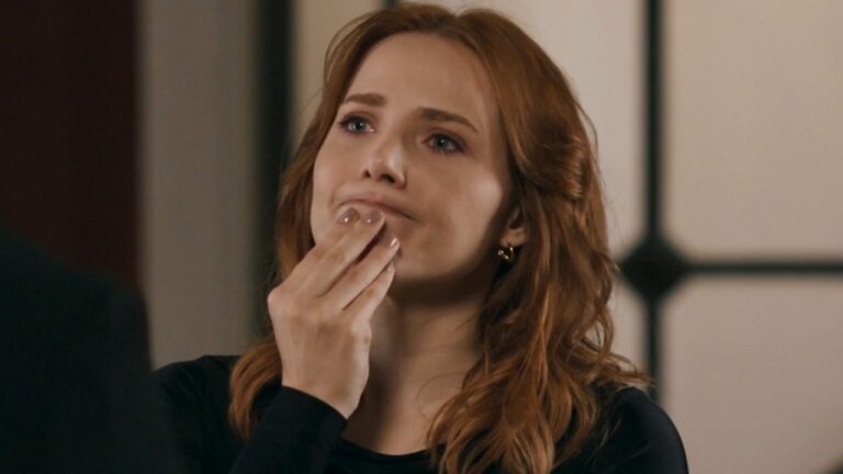 Adriana (Leticia Colin) aparece em primeiro plano, com cabelos ruivos soltos e blusa preta, enquanto leva a mão ao rosto e mantém uma expressão pensativa em cena da novela Quem Ama Cuida