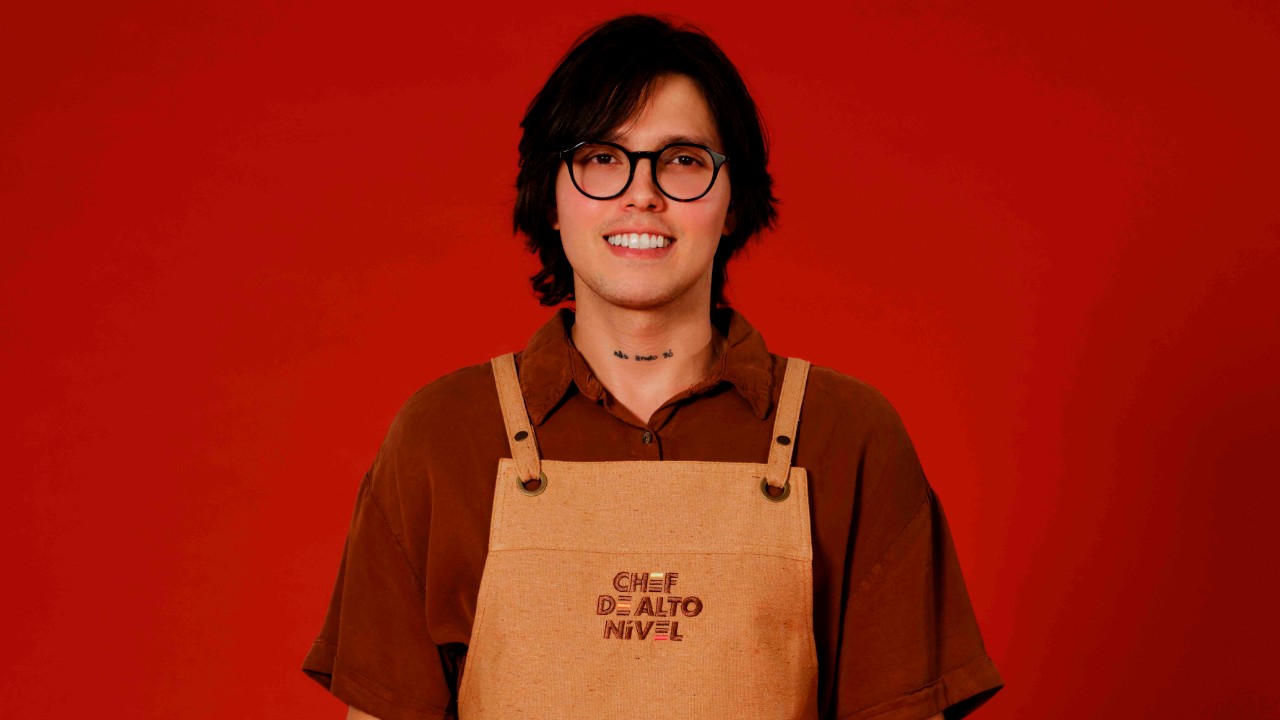 Alexandre Afonso sorri com óculos pretos, camisa marrom e avental bege de Chef de Alto Nível, diante de um fundo vermelho